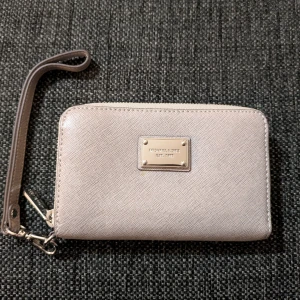 plånbok/korthållare/liten clutch från Michael Kors - Snygg grå plånbok från Michael Kors i saffiano-skinn med silverfärgad logoplatta framtill. Plånboken har dragkedja runtom, flera kortfack och ett handledsband. Perfekt för att hålla koll på dina kort och cash med stil. Köpt på Nordstrom i USA.