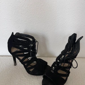 Svarta mocka pumps med snörning - Snygga svarta pumps från Gardenia i mocka med höga klackar och öppen tå. Skorna har coola utskurna detaljer och snörning framtill som ger en edgy vibe. Perfekta för dig som gillar att sticka ut med din stil.