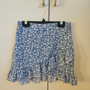 Blå blommig volangkjol med dragkedja. - Supersöt blå kortkjol med vitt blommönster och volangkant. Kjolen har en lätt A-linjeform och stängs med en diskret dragkedja i sidan. I storlek.. Perfekt för dig som gillar en feminin och somrig stil.