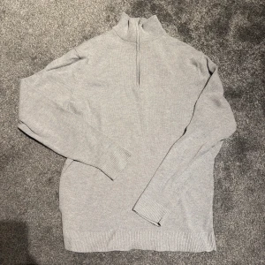 Ljusgrå stickad half zip-tröja - Snygg ljusgrå stickad tröja med half zip och hög krage. Tröjan har lång ärm och ribbade muddar vid ärmslut och nederkant. Perfekt att bära över en skjorta för en clean och stilren look. 