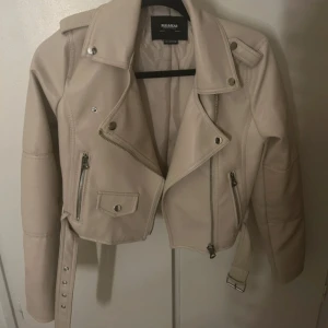 Beige skinnjacka från Pull&Bear - Snygg beige skinnjacka från Pull&Bear med croppad passform, klassisk bikerkrage, dragkedjor, knappar och bälte i midjan. Aldrig använd är i helt nytt skick
