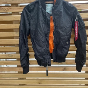 Svart bomberjacka från Alpha Industries - Säljer en svart bomberjacka från Alpha Industries med orange foder och klassisk röd tag på ärmen. Jackan har dragkedja framtill, ribbade muddar och fickor med tryckknapp. Perfekt för dig som vill ha en ikonisk streetstil.