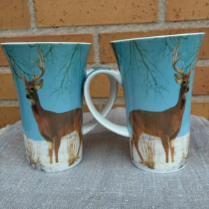 Keramiska muggar med hjortmotiv - Två keramikmuggar med vackert hjortmotiv mot blå bakgrund. Muggarna är ca 10 cm i diameter och har ett vinterlandskap. Fint skick utan synliga sprickor eller nagg, perfekt för kaffe eller te. Dekorativa och unika för naturälskare.