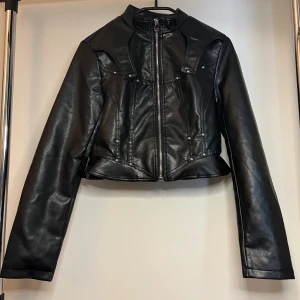Svart croppad skinnjacka med dragkedja - Snygg svart croppad skinnjacka med coola sömdetaljer och silvriga nitar. Jackan har hög krage, långa ärmar och dragkedja framtill. Perfekt för dig som vill ha en edgy look med lite biker-vibe.
