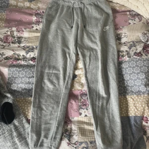 Grå mjukisbyxor från Nike - Gråa mjukisbyxor från Nike med broderad logga på vänster ben. Byxorna har resår och snörning i midjan samt muddar vid bensluten. Tillverkade i mjukt bomullsmaterial för extra komfort. Perfekta för chill eller träning.