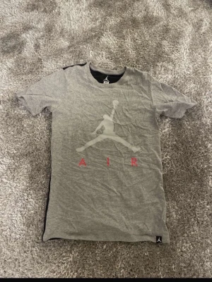 Grå och svart Jordan träningströja - Snygg t-shirt från Jordan i storlek XS. Framsidan är grå med stort Jumpman-tryck och röd AIR-text, medan baksidan är helsvart. Mjuk bomullskvalitet och korta ärmar. Perfekt för dig som gillar sportig streetwear och vill ha en ikonisk look.