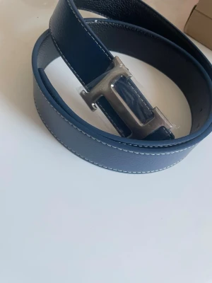 Mörkblå skinnbälte med H-spänne - Säljer ett helt nytt och oanvänt stilrent mörkblått bälte i skinn och svart skinnig sida som du kan byta sida med en stor, silverfärgad H-formad metallspänne. Bältet har vita sömmar längs kanterna och en klassisk, clean look som passar till jeans eller kostymbyxor.