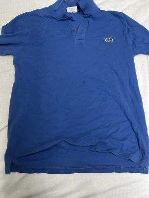 Blå pikétopp från Lacoste - Säljer en klassisk blå pikétopp från Lacoste med långa ärmar och krage. Toppen har knappar framtill och den ikoniska krokodilloggan broderad på bröstet. Perfekt för dig som gillar sportig och stilren look.