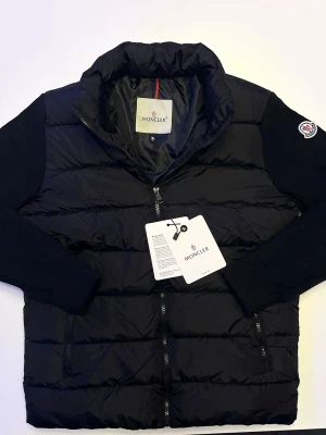 Svart monclair cardigan  - Säljer en svart pufferjacka från Moncler med stickade ärmar och klassisk Moncler-logga på armen. Jackan har dragkedja framtill och två sidofickor. Insidan har ett unikt Moncler-serietryck. Perfekt för dig som vill ha en stilren och varm jacka med exklusiv känsla.