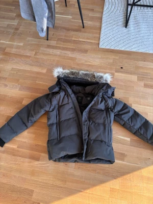 Svart pufferjacka från Canada Goose - Säljer en svart pufferjacka från Canada Goose i storlek L. Jackan har äkta päls på huvan, quiltad design och långa ärmar med muddar. Flera fickor och både dragkedja och knappar framtill. Perfekt för kalla dagar och riktigt snygg stil.
