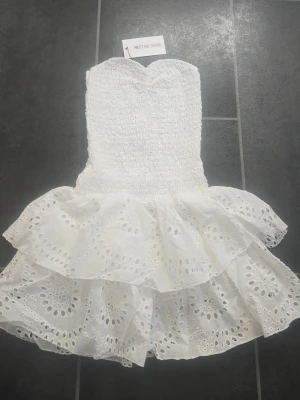 Vit axelbandslös kortklänning - Säljer en söt vit axelbandslös kortklänning från Meet Me There. Klänningen har smockad överdel och två lager volangkjol med broderade hålmönster. Perfekt för varma dagar. Helt ny- 