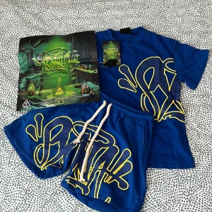 Syna world set. Blå och gul - Snygg blå t-shirt med stort gult graffiti-inspirerat tryck på både fram- och baksidan. T-shirten har klassisk passform och korta ärmar. Perfekt för dig som gillar streetwear och vill sticka ut med en färgstark look.