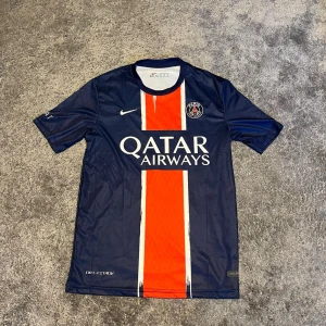 PSG tröja Dembele #10 24/25 - Paris Saint-Germain matchtröja från Nike med Dembele och nummer 10 på ryggen. Marinblå med röd och vit randig detalj framtill, PSG-logga på bröstet och Qatar Airways tryck. Materialet är polyester och har Dri-FIT teknologi. Ligue 1-märke på ärmen.