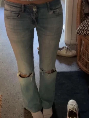 Ljusblå bootcut jeans med slitningar - Säljer ett par ljusblå jeans från nelly med bootcut-modell och slitna hål över knäna. Jeansen har klassiska fem fickor, knapp- och dragkedjegylf samt snygga knappar på bakfickorna.💕 tar emot bud💕