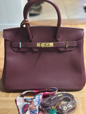 Vinröd handväska Genuine Leather - Säljer en lyxig vinröd handväska  i grained skinn med guldfärgade metalldetaljer. Väskan har klassisk Birkin-form, dubbla handtag och medföljande axelrem, lås och färgglad scarf. Insidan har dragkedjefack . Genuine leather