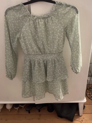 Grön blommig volangklänning KappAhl - Supersöt ljusgrön klänning från KappAhl med små vita blommor, volanger och smock i midjan. Klänningen har långa ärmar med resår vid ärmslut och en rundad halsringning med liten volangkant. Perfekt för vår och sommar! Använd 1 gång!