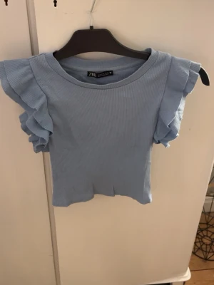 Ljusblå ribbad topp från Zara - Säljer en ljusblå ribbad topp från Zara i storlek S. Toppen har korta ärmar med volangdetaljer och rund halsringning. Materialet är mjukt och stretchigt, perfekt för en chill och snygg look. Passar dig som gillar stilrena och feminina detaljer.