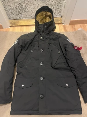 Svart parkajacka Alpha Industries S - Parkajacka från Alpha Industries i svart med snygga patchar på ärmarna, fuskpäls i huvan och flera fickor med både knappar och dragkedja. Jackan har långa ärmar, snörning i huvan och coola detaljer som röd tag och broderade märken. Perfekt för kalla dagar! Den är för DAM. Den är köpt för ca 3000kr från Zalando helt ny.
