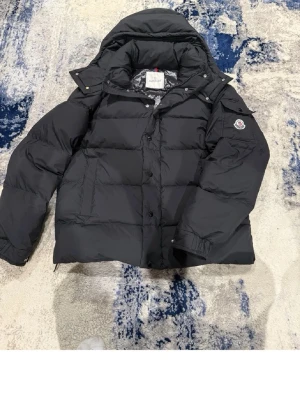 Svart Moncler pufferjacka med huva - Säljer en svart pufferjacka från Moncler i storlek L med huva och klassisk logga på ärmen. Jackan har knappar och dragkedja framtill, samt stora fickor. Perfekt för kalla dagar och riktigt snygg streetstil.