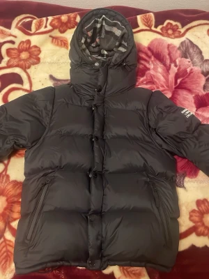 Svart Burberry pufferjacka med huva och väst  - Säljer en svart pufferjacka från Burberry med stor huva och rutig insida i grått, vitt och rött. Jackan har dragkedjor på fickorna, knappar framtill och en patch med Burberry London England på ärmen. Perfekt för kalla dagar och riktigt snygg streetstil.