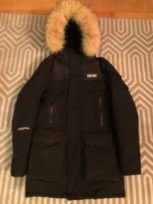 Svart jacka med päls - Svart parkajacka från FSBN med stor huva och beige fuskpäls. Jackan har flera fickor med lock och dragkedja, samt tryckt text på bröstet och ärmen. Perfekt för kalla dagar och har en riktigt schysst streetstil. Passar perfekt för storlek M.