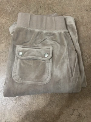 Beige mjukisbyxor Juicy Couture XS - Säljer ett par beige mjukisbyxor från Juicy Couture i storlek XS. Byxorna har bred resår i midjan, mjukt sammetstyg och en ficka med tryckknappar bak. Perfekta för chill och comfy vibes.