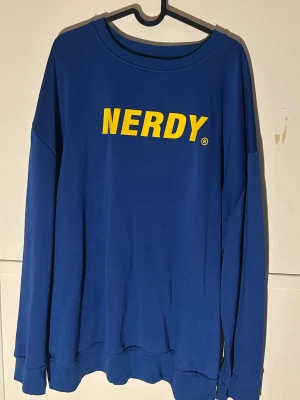 Blå sweatshirt från NERDY - Säljer en blå sweatshirt från NERDY med gul text framtill. Tröjan har en avslappnad passform och långa ärmar. Perfekt för dig som gillar streetwear och vill sticka ut med en färgglad look.