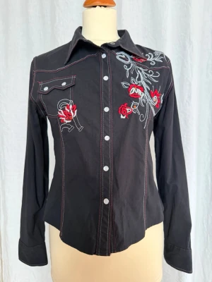 Svart broderad cowboyskjorta strl 38 - Svart skjorta med lång ärm och snygga röda och vita broderier med blommor och detaljer på både fram- och baksida. Skjortan har vita tryckknappar, röd kontrastsöm och klassisk krage. Materialet är bomull och passformen är normal. Finns inte i Sverige, inköpt utomlands i början av 2000. 