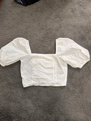 Vit blus med puffärmar och broderi - Säljer en vit blus med fyrkantig urringning och puffiga kortärmar med broderade detaljer. Toppen är croppad och har smock i ryggen för skön passform. Materialet är bomull och är lätt och luftigt, perfekt för varmare dagar. Finns en liten fläck på tröjan.