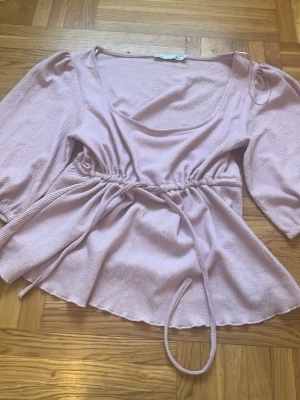 Ljusrosa omlottblus med knyt - Säljer en ljusrosa blus med omlottdesign och knyt i midjan. Blusen har trekvartsärmar och är tillverkad i ett mjukt, strukturerat tyg som ger en skön känsla. Perfekt för dig som gillar feminina detaljer och en avslappnad passform.