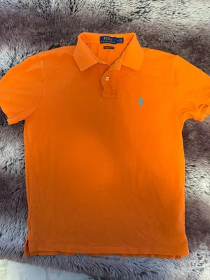Orange pikétröja från Polo Ralph Lauren - Säljer en orange pikétröja från Polo Ralph Lauren i modellen Custom Slim Fit. Tröjan har korta ärmar, klassisk krage med knappslå och den ikoniska blå broderade loggan på bröstet. Tillverkad i bomull.