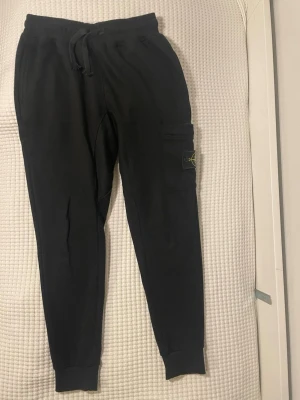 Svarta mjukisbyxor från Stone Island - Svarta mjukisbyxor från Stone Island med klassisk logga på benfickan. Byxorna har snörning i midjan, ribbade muddar vid benslut och flera fickor med knappstängning. Sköna och stilrena, perfekta för chill eller streetwear. Materialet är mjukt bomullstyg.