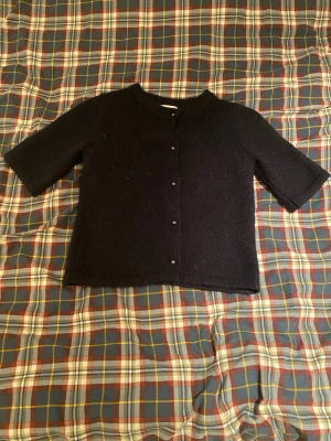 Svart kofta från Gina Tricot - Mysig svart kofta från Gina Tricot med korta ärmar och rund hals. Koftan har knappar framtill och är stickad i ett mjukt material som passar perfekt för lager-på-lager. Enkel och stilren design som funkar till många olika looks.