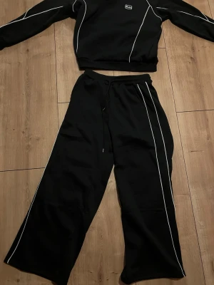 Svart mjukisset med vita detaljer - Svartset med vita kontrastsömmar längs benen. Byxorna har dragsko i midjan och en loose, relaxed passform. Tillverkade i mjukt material som känns skönt mot huden. 