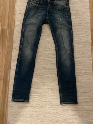 Replay Anbass jeans mörkblå 28/32 - Snygga Replay Anbass jeans i mörkblå tvätt med slitningar och klassisk femficksdesign. Nypris 1900kr. Säljer pga att jag inte använder dom längre Modellen är slim fit med smal passform och raka ben. Jeansen har detaljerade sömmar och läderpatch bak i midjan. Perfekta för dig som gillar en modern och stilren look.