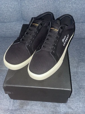 Svarta Saint Laurent sneakers - Snygga svarta sneakers från Saint Laurent med vit sula och diskret logga på sidan. Skorna har rund tå, snörning och är tillverkade i textil och skinn. Perfekt för dig som gillar stilrena och exklusiva sneakers med en clean look.