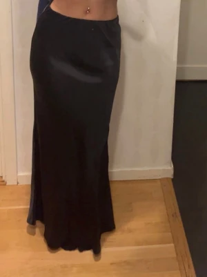 Svart satin maxikjol strl 32 - Säljer en svart långkjol i satin med snyggt åtsittande passform och lätt utsvängd nederdel. 