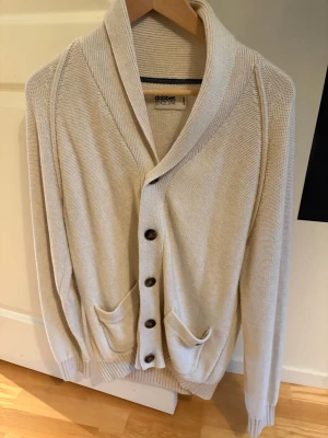 Beige kofta från Dobber med knappar - Säljer en beige stilren kofta från Dobber med grovstickad struktur och bred krage. Koftan har fyra mörka knappar framtill, två fickor och ribbade muddar vid ärmslut och nederkant. 