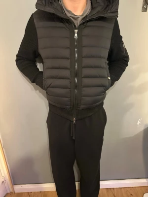Svart Moncler pufferjacka med stickade ärmar - Svart pufferjacka från Moncler med stickade ärmar. Jackan har dragkedja framtill, sidofickor med knapp och en patch med Moncler-logga på ärmen. Kroppen är quiltad och ärmarna är ribbstickade för en schysst kontrast. Jackan är helt utan några skavanker.