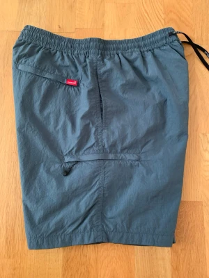  Levi's badbyxor  - Säljer ett par gråa badbyxor från Levi's med elastisk midja och snörning. De har en enkel design med praktiska fickor, varav en med dragkedja. Levi's-loggan syns på bakfickan.De är tillverkade i lätt polyamid som torkar snabbt.