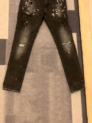 Svarta skinny jeans med slitningar - Säljer ett par svarta jeans med skinny fit och coola slitningar och hål på både fram- och baksida. Jeansen har en smal passform och är tillverkade i klassiskt denim-material. Perfekta för dig som gillar en edgy streetstil.