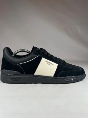 Valentino Garavani sneakers svart/vit - Svarta och vita sneakers från Valentino Garavani med snygg kontrastpanel i vitt på sidan och logotyp. Skorna har snörning, platt sula med nitar och är tillverkade i mocka och skinn. Perfekt för dig som gillar stilrena och lyxiga detaljer. Bara använda ett fåtal gånger och tagit bra hand om dom men tyvärr är det inte riktigt min stil