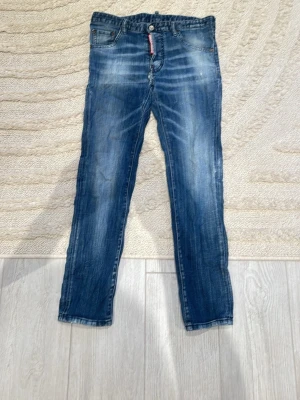 Blå jeans från Dsquared2 - Säljer ett par snygga blå jeans från Dsquared2 med klassisk femficksdesign och läderpatch bak. Jeansen har en normal passform, raka ben och coola slitningar framtill. Materialet är jeans i bomull och färgen är en tvättad blå med ljusare partier. Jeansen har ett litet håll men man kan sy det lätt som en plätt