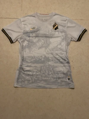 AIK x Nike vit fotbollströja XL - Säljer en riktigt snygg AIK fotbollströja från Nike i vitt med grått tryck av Stockholmsmotiv. Tröjan har svarta och gula detaljer, AIK-emblem på bröstet och Nike-logga i guld. Tillverkad i Dri-FIT material som andas. Perfekt för match eller samling.