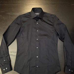 Svart slim fit skjorta Dressmann XS - Snygg svart skjorta från Dressmann i slim fit-modell. Tillverkad i premium bomull med stretch för extra komfort. Klassisk krage och knappar framtill, perfekt för dig som gillar stilrena och enkla plagg.