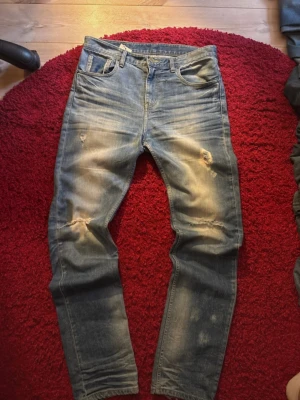 Blå jeans med slitningar  - Säljer ett par blå jeans med coola slitningar och hål på knäna. Jeansen har klassisk femficksdesign, ljus tvätt och en snygg patch med text bak i midjan. Passformen är rak och materialet är robust denim. Perfekt för dig som gillar en avslappnad streetstil.