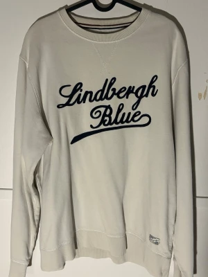 Vit sweatshirt Lindbergh Blue XL - Vit sweatshirt från Lindbergh Blue i storlek XL med mörkblå broderad text på bröstet. Tröjan har rund hals, ribbade muddar och är långärmad. Materialet är mjukt och känns som bomull, perfekt för chill dagar. Litet hål i ena armhålan.
