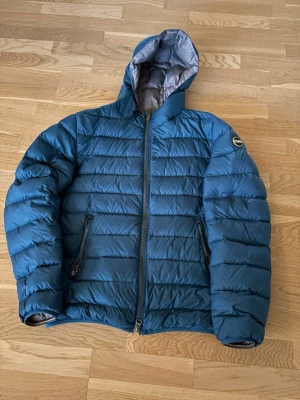 Blå pufferjacka från Colmar - Säljer en blå pufferjacka från Colmar Originals med huva och två dragkedjeförsedda fickor. Jackan har horisontella quiltade partier och en snygg, sportig look. Perfekt för kyliga dagar och enkel att matcha med olika outfits. Kan tänka mig att gå ner i pris.