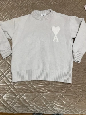 Ljusgrå stickad tröja från Ami - Säljer en ljusgrå stickad tröja från Ami Alexandre Mattiussi med ett stort vitt hjärta och bokstaven A broderat på bröstet. Tröjan har rund hals, ribbade muddar och är långärmad. Perfekt för dig som gillar stilrena detaljer och mjuka material. Kommer med påse och tag 