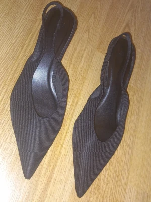 Svarta pumps med spetsig tå - Säljer ett par svarta pumps med spetsig tå och låg klack. Skorna har en elegant och smal siluett med öppen häl och elastiskt band bak. Dessa är endast använda en gång.